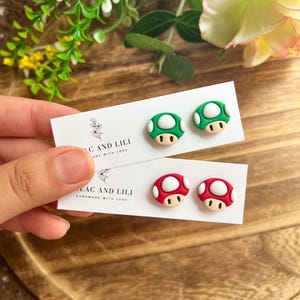 May include: A pair of white cards with mushroom-shaped earrings. One card has green and white mushroom earrings, and the other has red and white mushroom earrings. The cards read "LAC AND LILI HANDMADE WITH LOVE."