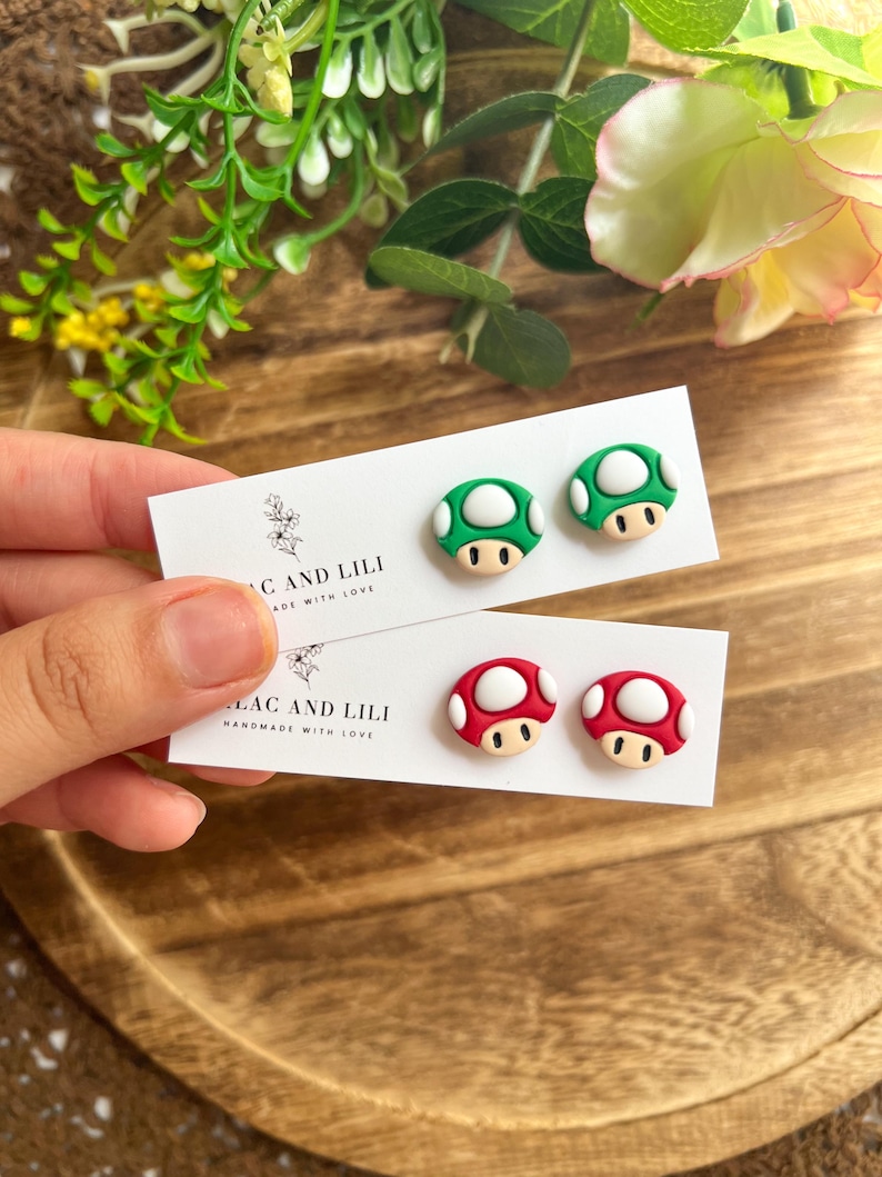 May include: A pair of white cards with mushroom-shaped earrings. One card has green and white mushroom earrings, and the other has red and white mushroom earrings. The cards read "LAC AND LILI HANDMADE WITH LOVE."