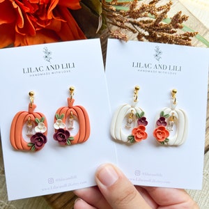 May include: Two pairs of pumpkin-shaped earrings with floral accents. The earrings are made of polymer clay and have a white and orange color scheme. The earrings are handmade and have the text "Lilac and Lili Handmade with Love" on the cards they are displayed on.
