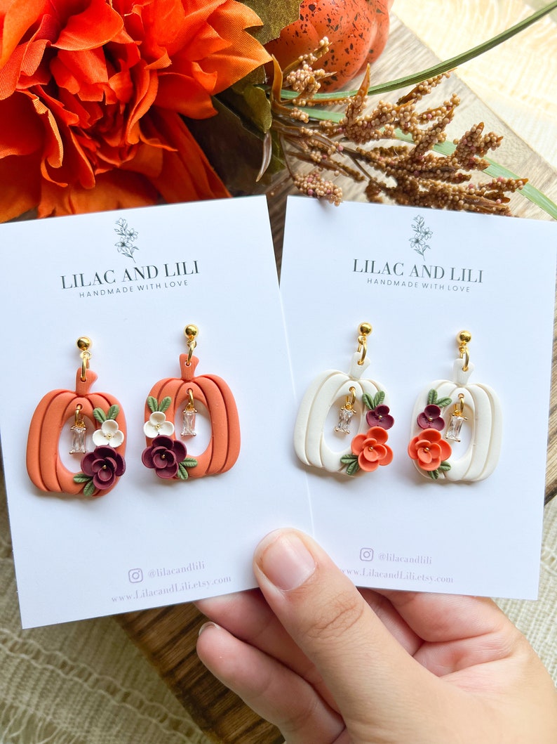 May include: Two pairs of pumpkin-shaped earrings with floral accents. The earrings are made of polymer clay and have a white and orange color scheme. The earrings are handmade and have the text "Lilac and Lili Handmade with Love" on the cards they are displayed on.