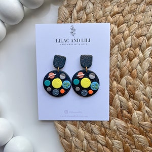 May include: A pair of black and gold earrings with a galaxy theme. The earrings are made of polymer clay and feature a variety of colours, including blue, green, yellow, and orange. The earrings are handmade with love by Lilac and Lili.