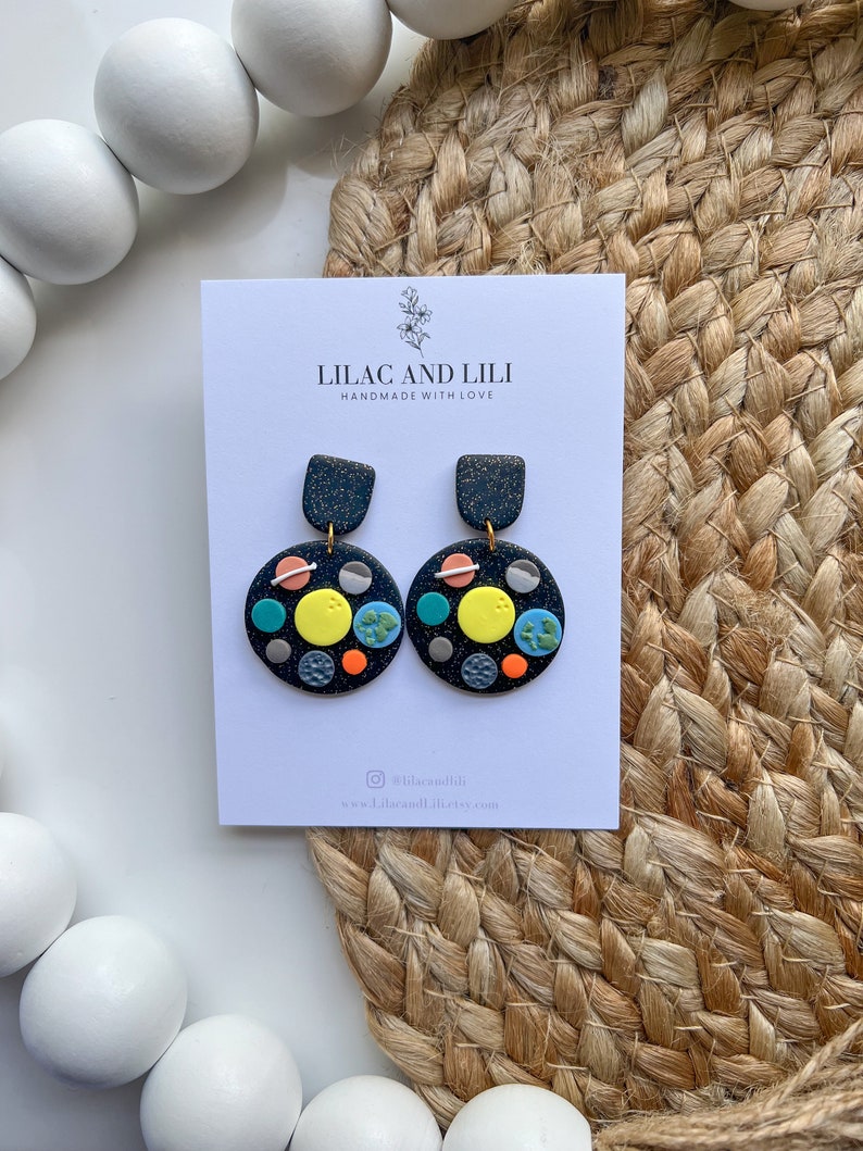 May include: A pair of black and gold earrings with a galaxy theme. The earrings are made of polymer clay and feature a variety of colours, including blue, green, yellow, and orange. The earrings are handmade with love by Lilac and Lili.