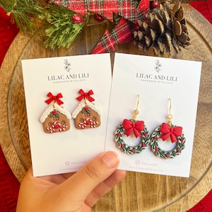 May include: Two pairs of Christmas earrings on white cards. One pair features gingerbread house designs with red bows and candy canes. The other pair has green wreath designs with red bows. The cards read "LILAC AND LILI HANDMADE WITH LOVE."
