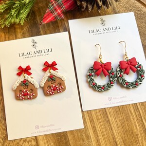 May include: Two pairs of festive earrings on white cards. One pair features gingerbread house designs with red bows and candy canes. The other pair showcases wreath designs with red bows and green leaves. The cards read "LILAC AND LILI HANDMADE WITH LOVE".