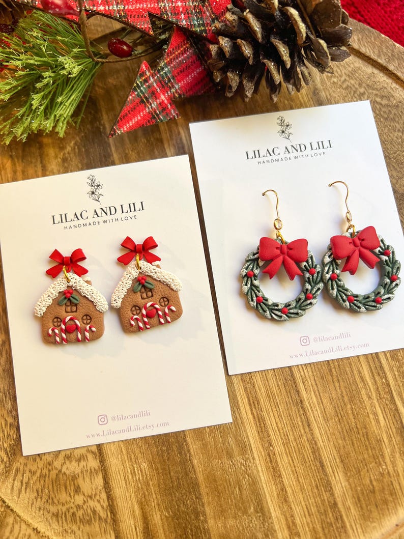 May include: Two pairs of festive earrings on white cards. One pair features gingerbread house designs with red bows and candy canes. The other pair showcases wreath designs with red bows and green leaves. The cards read "LILAC AND LILI HANDMADE WITH LOVE".
