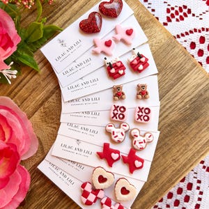May include: A collection of Valentine's Day-themed stud earrings in various designs, including hearts, Texas shapes, teddy bears, and "XO" text. The earrings are red, pink, and white, and are displayed on small cards with the text "LILAC AND LILI HANDMADE WITH LOVE".