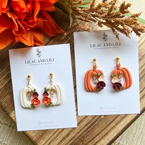 May include: Two pairs of handmade polymer clay pumpkin earrings. One pair is white with orange and burgundy flowers and the other pair is orange with white and burgundy flowers. Both pairs have gold ear wires and clear rhinestones.
