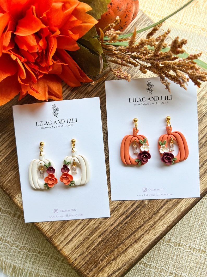 May include: Two pairs of handmade polymer clay pumpkin earrings. One pair is white with orange and burgundy flowers and the other pair is orange with white and burgundy flowers. Both pairs have gold ear wires and clear rhinestones.