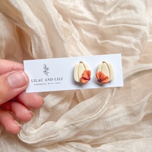 May include: A pair of cream-colored earrings with a small orange flower on each. The earrings are on a white card with the text "Lilac and Lili Handmade with Love".
