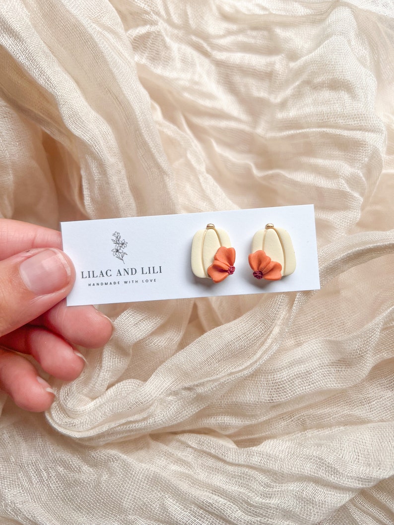 May include: A pair of cream-colored earrings with a small orange flower on each. The earrings are on a white card with the text "Lilac and Lili Handmade with Love".