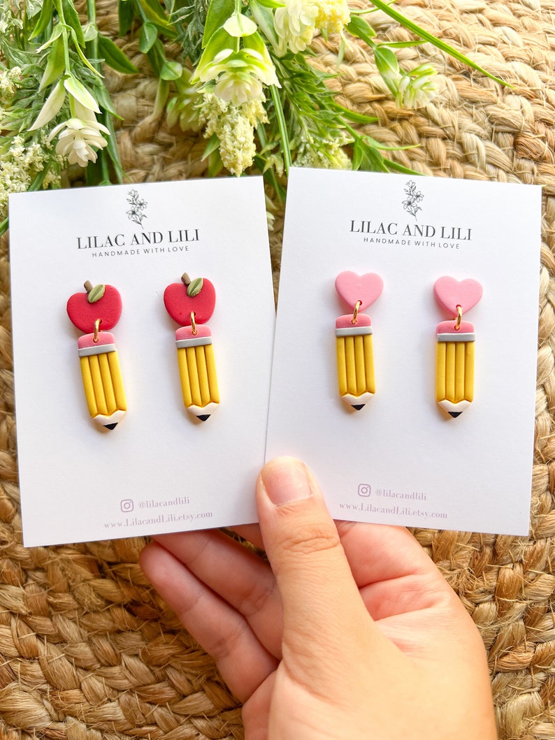 Pu&ograve; includere: Due paia di orecchini fatti a mano in pasta polimerica. Il primo paio presenta una matita gialla con una mela rossa in cima. Il secondo paio presenta una matita gialla con un cuore rosa in cima. Entrambe le paia hanno uno sfondo bianco con il testo "Lilac and Lili Handmade with Love" e informazioni sui social media.