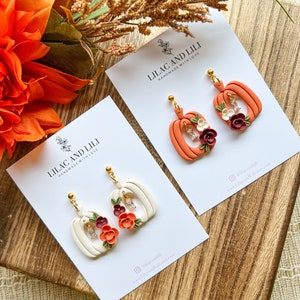 May include: Two pairs of pumpkin-shaped earrings with floral accents. The earrings are made of polymer clay and have a white and orange color scheme. The earrings are handmade with love by Lilac and Lili.