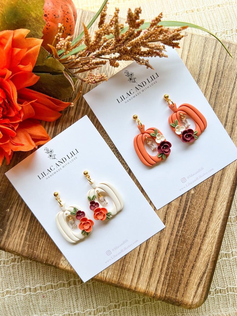 May include: Two pairs of pumpkin-shaped earrings with floral accents. The earrings are made of polymer clay and have a white and orange color scheme. The earrings are handmade with love by Lilac and Lili.