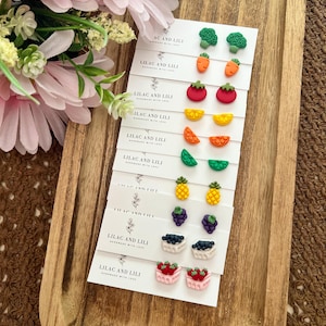May include: A collection of nine pairs of miniature food-themed stud earrings attached to white display cards labeled Lilac and Lili. The designs include broccoli, carrots, tomatoes, citrus slices, pineapples, grapes, and baskets of berries.