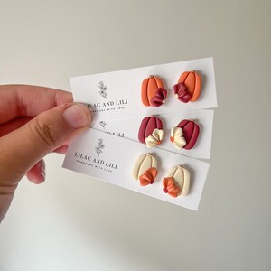 May include: Three pairs of polymer clay pumpkin earrings with different colored flowers. The pumpkins are orange, white, and red. The flowers are burgundy, cream, and orange. The earrings are handmade by Lilac and Lili.