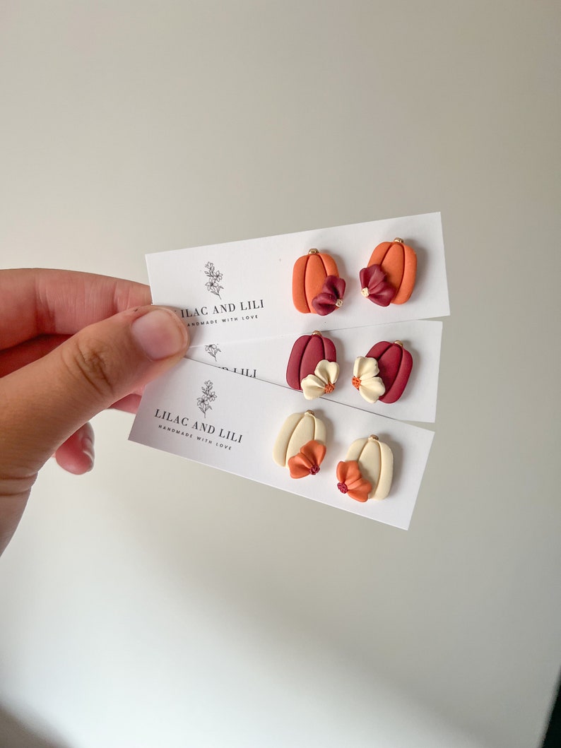 May include: Three pairs of polymer clay pumpkin earrings with different colored flowers. The pumpkins are orange, white, and red. The flowers are burgundy, cream, and orange. The earrings are handmade by Lilac and Lili.