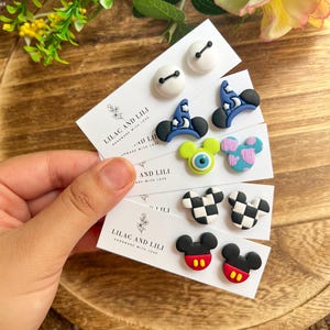 May include: A variety of handmade stud earrings featuring cartoon designs. The earrings include white button shapes, blue wizard hats, green monster eyes, pink and blue spotted shapes, black and white checkered shapes, and Mickey Mouse designs. The cards read "LILAC AND LILI".