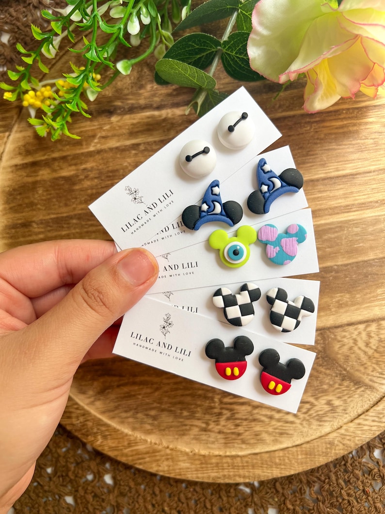 May include: A variety of handmade stud earrings featuring cartoon designs. The earrings include white button shapes, blue wizard hats, green monster eyes, pink and blue spotted shapes, black and white checkered shapes, and Mickey Mouse designs. The cards read "LILAC AND LILI".