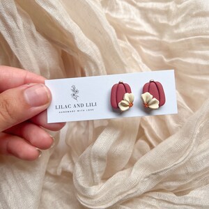 May include: A pair of red pumpkin-shaped earrings with a white flower accent. The earrings are handmade and the card reads "Lilac and Lili Handmade with Love."