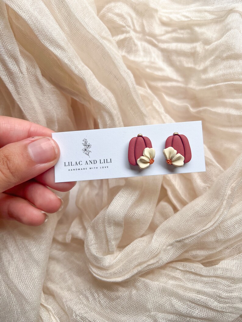 May include: A pair of red pumpkin-shaped earrings with a white flower accent. The earrings are handmade and the card reads "Lilac and Lili Handmade with Love."