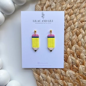 May include: A pair of yellow pencil earrings with pink eraser details and gold accents. The earrings are handmade and feature a simple design. The earrings are displayed on a white card with the text "Lilac and Lili Handmade with Love" and the social media handle "@lilacandili".