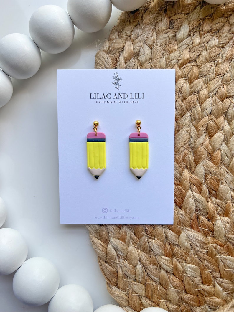 May include: A pair of yellow pencil earrings with pink eraser details and gold accents. The earrings are handmade and feature a simple design. The earrings are displayed on a white card with the text "Lilac and Lili Handmade with Love" and the social media handle "@lilacandili".