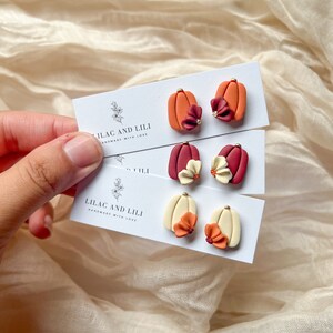 May include: Three pairs of polymer clay pumpkin earrings with flower accents. The earrings are displayed on white cards with the text "Lilac and Lili Handmade with Love".