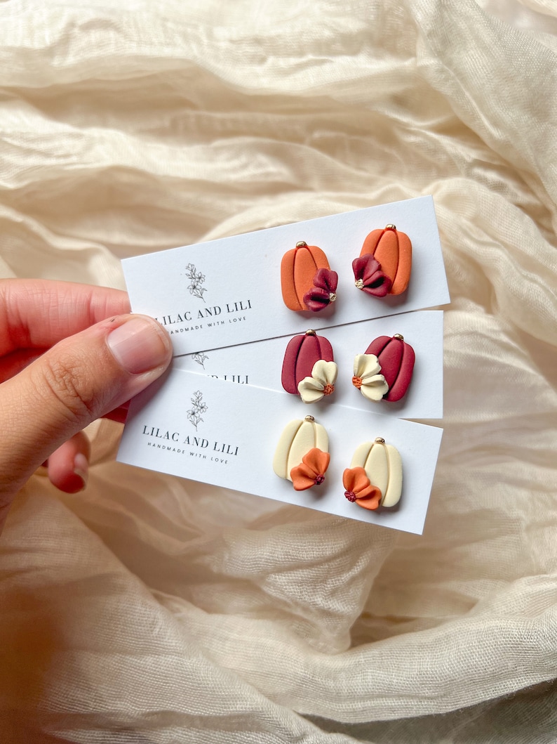 May include: Three pairs of polymer clay pumpkin earrings with flower accents. The earrings are displayed on white cards with the text "Lilac and Lili Handmade with Love".