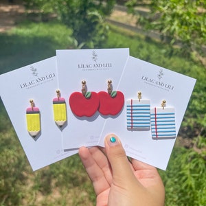 May include: Three cards with the text "Lilac and Lili Handmade with Love" and images of earrings. The earrings are a pair of red apples, a pair of yellow pencils, and a pair of blue and white striped paper.