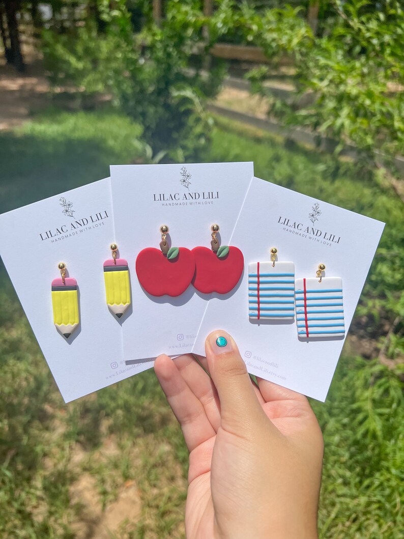 May include: Three cards with the text "Lilac and Lili Handmade with Love" and images of earrings. The earrings are a pair of red apples, a pair of yellow pencils, and a pair of blue and white striped paper.