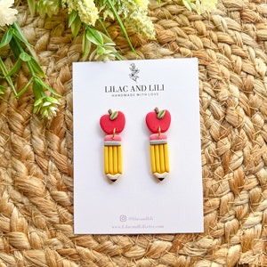 Pu&ograve; includere: Un paio di orecchini a forma di matita gialla con ciondoli a forma di mela rossa. Gli orecchini sono fatti a mano e hanno un dettaglio di gomma rosa. La carta dietro gli orecchini dice "Lilac and Lili Handmade with Love".