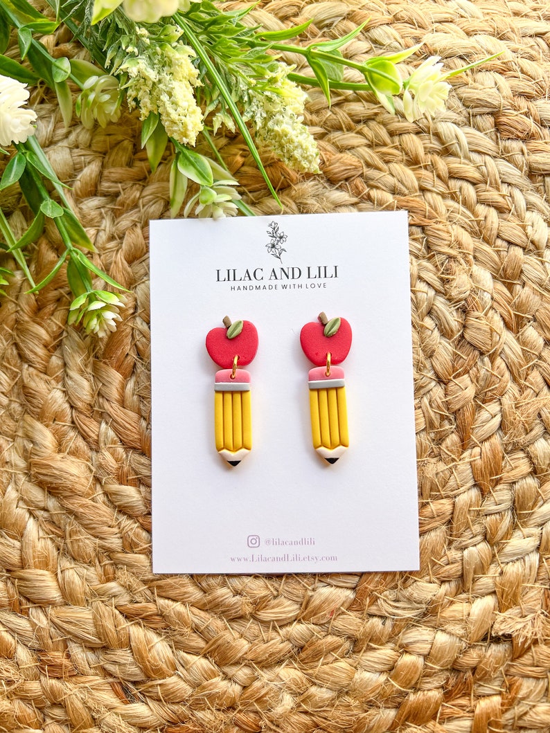 Pu&ograve; includere: Un paio di orecchini a forma di matita gialla con ciondoli a forma di mela rossa. Gli orecchini sono fatti a mano e hanno un dettaglio di gomma rosa. La carta dietro gli orecchini dice "Lilac and Lili Handmade with Love".