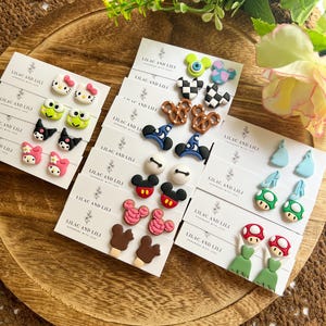 May include: A collection of handmade stud earrings featuring various designs, including cartoon characters, pretzels, and mushrooms. The earrings are displayed on small white cards with the text "LILAC AND LILI". The earrings are on a wooden tray.