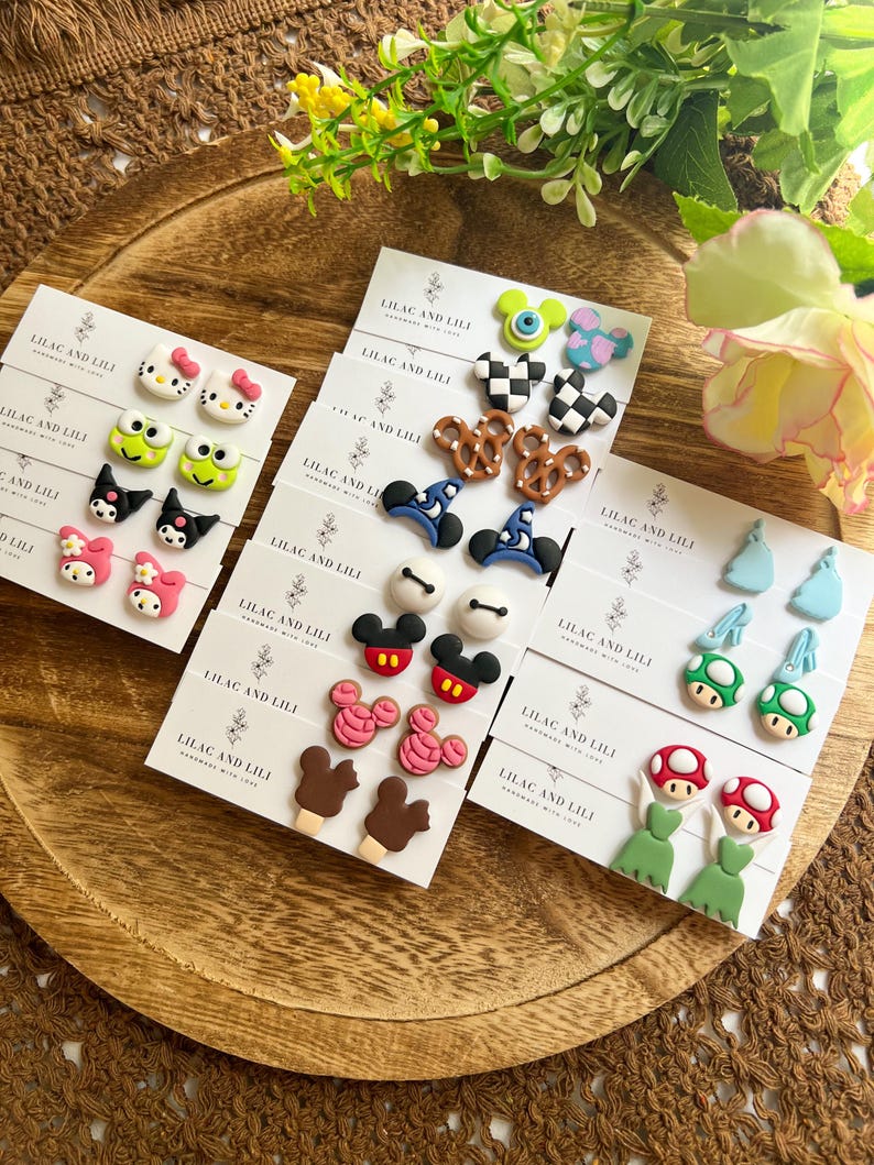 May include: A collection of handmade stud earrings featuring various designs, including cartoon characters, pretzels, and mushrooms. The earrings are displayed on small white cards with the text "LILAC AND LILI". The earrings are on a wooden tray.