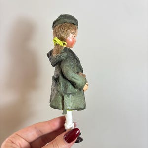 May include: A vintage doll figurine of a child wearing a green coat and hat. The doll has blonde hair with a yellow ribbon and is holding a small object. The doll is wearing white trousers and black shoes.