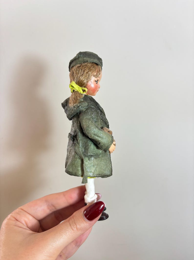 May include: A vintage doll figurine of a child wearing a green coat and hat. The doll has blonde hair with a yellow ribbon and is holding a small object. The doll is wearing white trousers and black shoes.