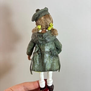 May include: A handcrafted doll, seen from behind, dressed in a green coat and beret. The doll features blonde hair with yellow bows, white socks, and brown shoes. The coat has a textured finish.