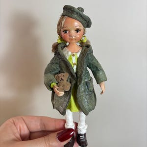 May include: A handmade doll wearing a green coat and beret, holding a small teddy bear. The doll has blonde hair, a lime green dress, white leggings, and brown shoes. The doll's face is painted with delicate features.