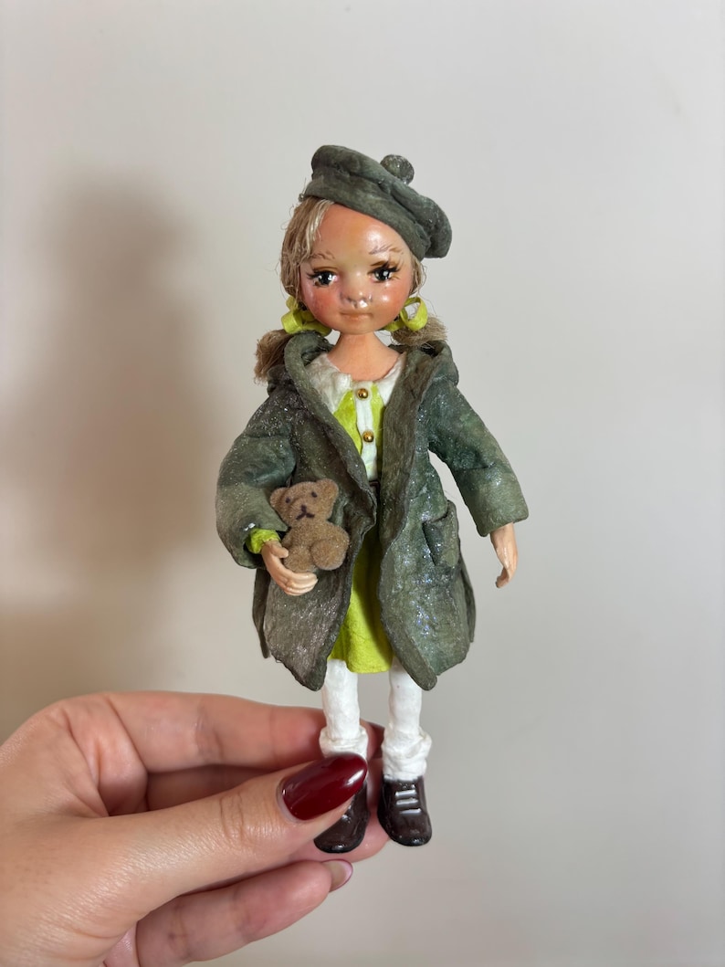 May include: A handmade doll wearing a green coat and beret, holding a small teddy bear. The doll has blonde hair, a lime green dress, white leggings, and brown shoes. The doll's face is painted with delicate features.