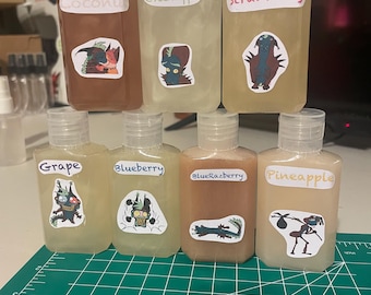 May include: A collection of eight clear plastic bottles filled with colored liquid, each labeled with a different fruit flavor: Coconut, Green Apple, Strawberry, Grape, Blueberry, Blue Razberry, and Pineapple. Each bottle has a unique cartoon sticker.