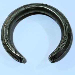 May include: A silver-toned cuff bracelet with an open design. The bracelet features etched geometric patterns and circular details along its surface. The metal has a slightly aged appearance, suggesting it may be vintage or antique.