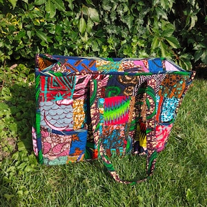 Op de afbeelding: Een kleurrijke patchwork tote bag met diverse patronen en kleuren, waaronder rood, blauw, groen en bruin. De tas heeft twee buitenzakken en een lange handgreep. De tas ligt op groen gras.