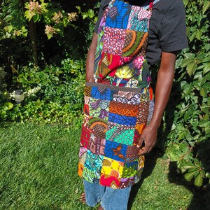 May include: A colorful patchwork apron with a variety of patterns and colors, including red, blue, green, and yellow. The apron has a large pocket and is worn by a person in blue jeans and a black t-shirt. The apron is made of fabric.