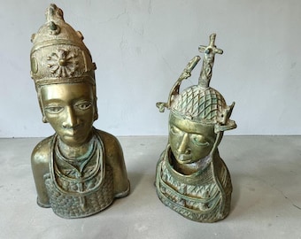 May include: Two metallic bronze-toned busts depicting human figures. The taller bust features a high, ornate headdress with a central circular emblem. The shorter bust wears a textured, grid-patterned helmet topped with a cross-like finial.