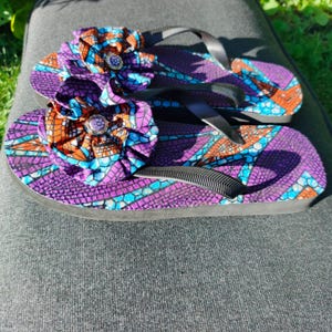 May include: A pair of flip-flops featuring a vibrant purple, orange, and blue geometric mosaic pattern on the footbed. Each sandal is adorned with a large, ruffled fabric flower in matching colors, centered with a small, circular purple embellishment.