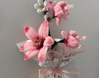 May include: A bouquet of handmade pink and white fabric flowers, including lilies and daisies, wrapped in paper and tied with a sheer pink ribbon. The bouquet is held in a hand.