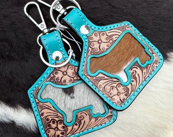 May include: Two leather keychains with a turquoise border and silver hardware. The keychains feature a cowhide design with a cow silhouette and floral tooling. The background is a black and white cowhide pattern.