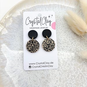 May include: A pair of black and beige dangle earrings with a leopard print design. The earrings are handmade and feature a black circle on top of a larger beige circle with a black leopard print.