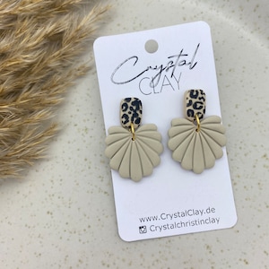Pode incluir: Um par de brincos de concha bege com um detalhe de estampa de leopardo preto e marrom. Os brincos são exibidos em um cartão branco com o texto "Crystal Clay" e informações do site.