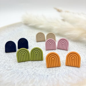 Puede incluir: Un conjunto de pendientes de botón con forma de arcoíris en varios colores, incluyendo beige, rosa, verde, azul y naranja. Los pendientes están hechos de arcilla polimérica y tienen un acabado mate.